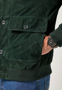 Giacca in suede verde con chiusura frontale a bottoni, ampie tasche sul petto, polsini e orlo a coste. Vestibilità slim con una superficie liscia e opaca.