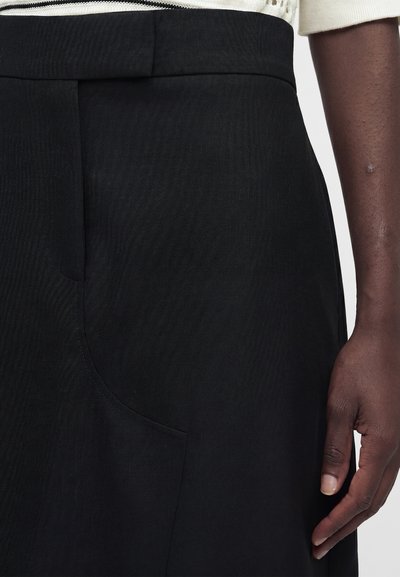 Κοντινό ενός ατόμου που φοράει μαύρο ψηλόμεσο παντελόνι με πλαϊνή τσέπη και λευκό πλεκτό τοπ, το χέρι ακουμπάει στο πλευρό.