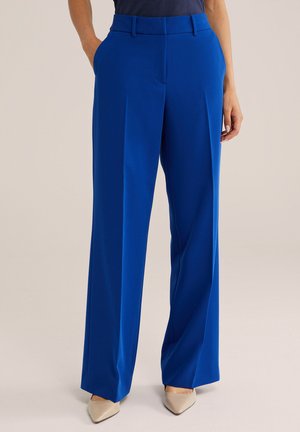 Femme portant un pantalon large bleu vif avec des poches latérales et des talons beiges à bout pointu sur un fond uni.