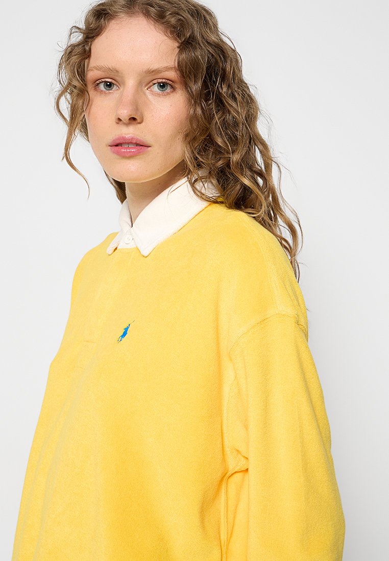 Felpa gialla con una texture morbida, caratterizzata da un'accattivante logo blu. Il design include una camicia bianca con colletto sotto.