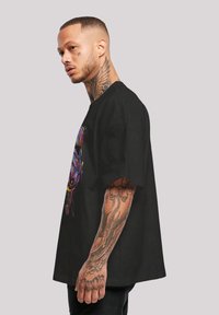 Juoda per didelė t-shirt maikė su spalvingu grafiniu dizainu. Trumpomis rankovėmis ir laisvu siluetu. Modelis demonstruoja šoną, matomi tatuiruotės.