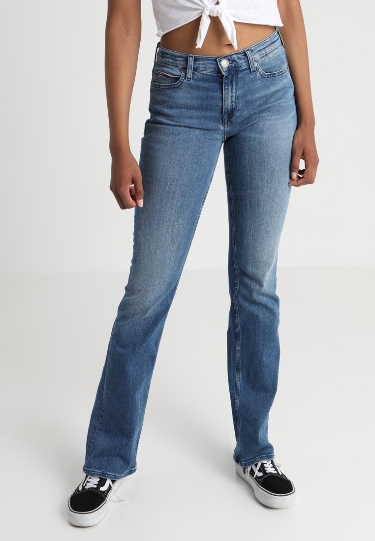 Mujer con vaqueros azules de talle medio, ligeramente acampanados, top corto blanco anudado y zapatillas blancas y negras, de pie frente a un fondo liso.