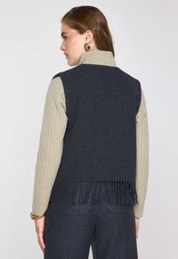 Donna di profilo che indossa orecchini a cerchio dorati, un dolcevita rigato beige, un gilet senza maniche con frange blu navy e pantaloni blu navy.