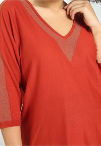 Rode gebreide top met een diepe V-hals en een doorschijnende, metalen accent. Korte mouwen en geribbelde manchetten voegen textuur toe aan het ontwerp.