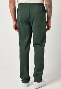 Pantalones jogger verdes con textura, cintura elástica y un bolsillo trasero, combinados con zapatillas blancas, vistos desde la parte de atrás.