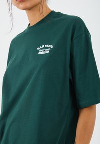 Green cotton t-shirt featuring a crew neck, short sleeves, and white printed text on the front. Soft texture with a relaxed fit.