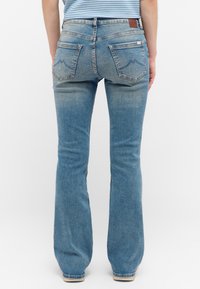 Vaqueros de mezclilla azul con un acabado desgastado, que presentan un parche rectangular de cuero en la cinturilla trasera y dos bolsillos traseros con detalles de costura.