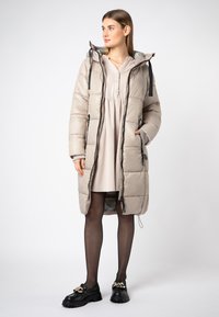 Gevoerde lange beige jas met een capuchon, voorklep met rits en een trekkoord. Gedragen over een lichtgekleurde jurk. Zwarte schoenen met een kettingaccent.