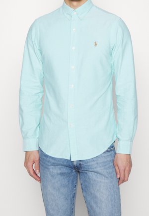 Mann trägt ein hellblaues Hemd mit Knöpfen und einem kleinen gestickten Logo auf der Brust, kombiniert mit hellblauen Jeans.