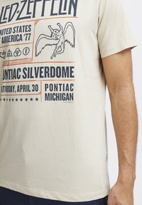 Beidža kokvilnas t-krekls ar melnu un oranžu drukātu tekstu un grafikām, ieskaitot spārnotu figūru un koncerta detaļas no Pontiac Silverdome.