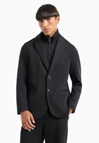 Blazer nero in tessuto elasticizzato, con colletto a risvolto, due tasche anteriori e due bottoni. Indossato sopra un top a collo alto.