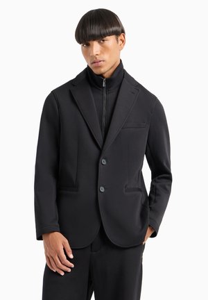 Blazer nero in tessuto elasticizzato, con colletto a risvolto, due tasche anteriori e due bottoni. Indossato sopra un top a collo alto.