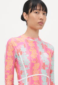 Rash guard à manches longues rose avec un motif floral en orange et turquoise, doté d'accents blancs et d'un logo à l'avant.