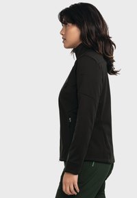 Schwarze Softshelljacke mit hohem Kragen, seitlichen Reißverschlusstaschen und taillierter Passform. Strukturierter Stoff für Wärme, kombiniert mit grünen Hosen.