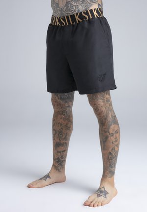 Mann mit tätowierten Beinen, der schwarze Shorts mit goldenem, gebrandetem elastischem Bund trägt und barfuß vor einem schlichten Hintergrund steht.