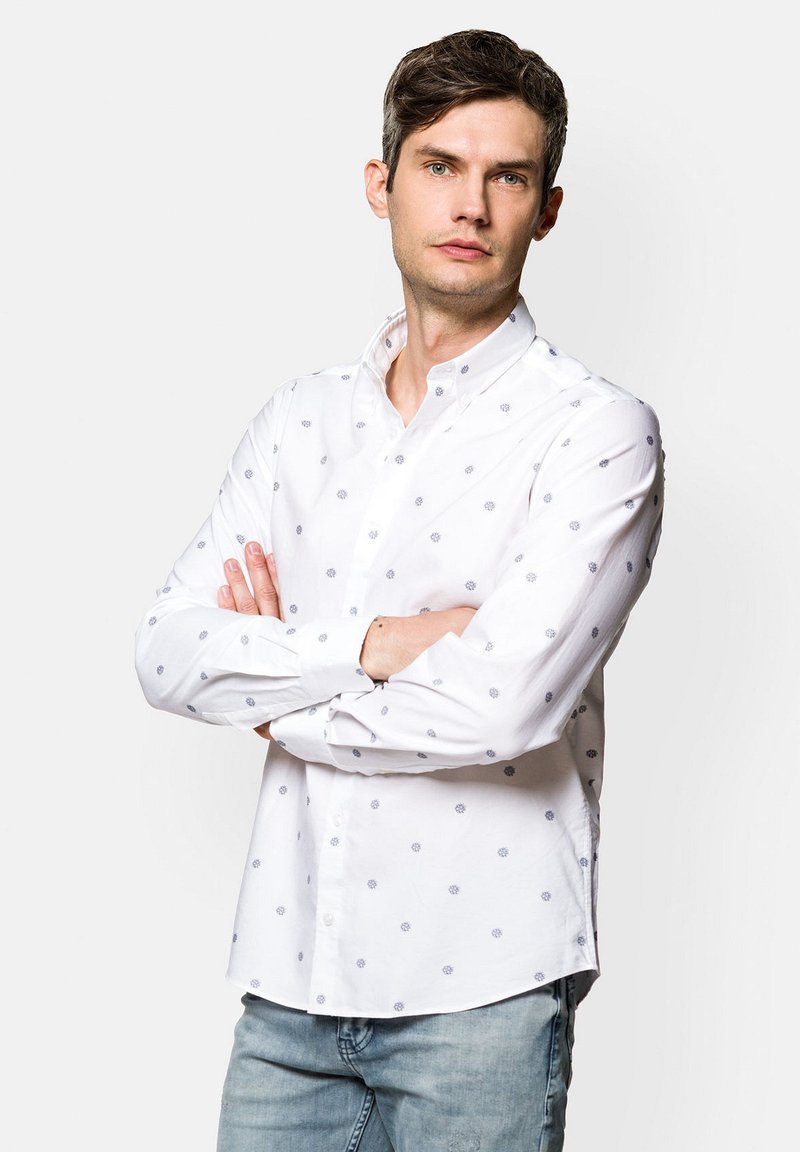 Biała koszula z długim rękawem, z niebieskimi wzorami kwiatowymi i kołnierzykiem, połączona z jasnoniebieskimi dżinsami.