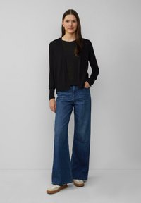 Zwarte open voorkaardigan over een donkere top, gecombineerd met wijde blauwe spijkerbroeken en beige sneakers met witte accenten. Soepele texturen door het geheel.