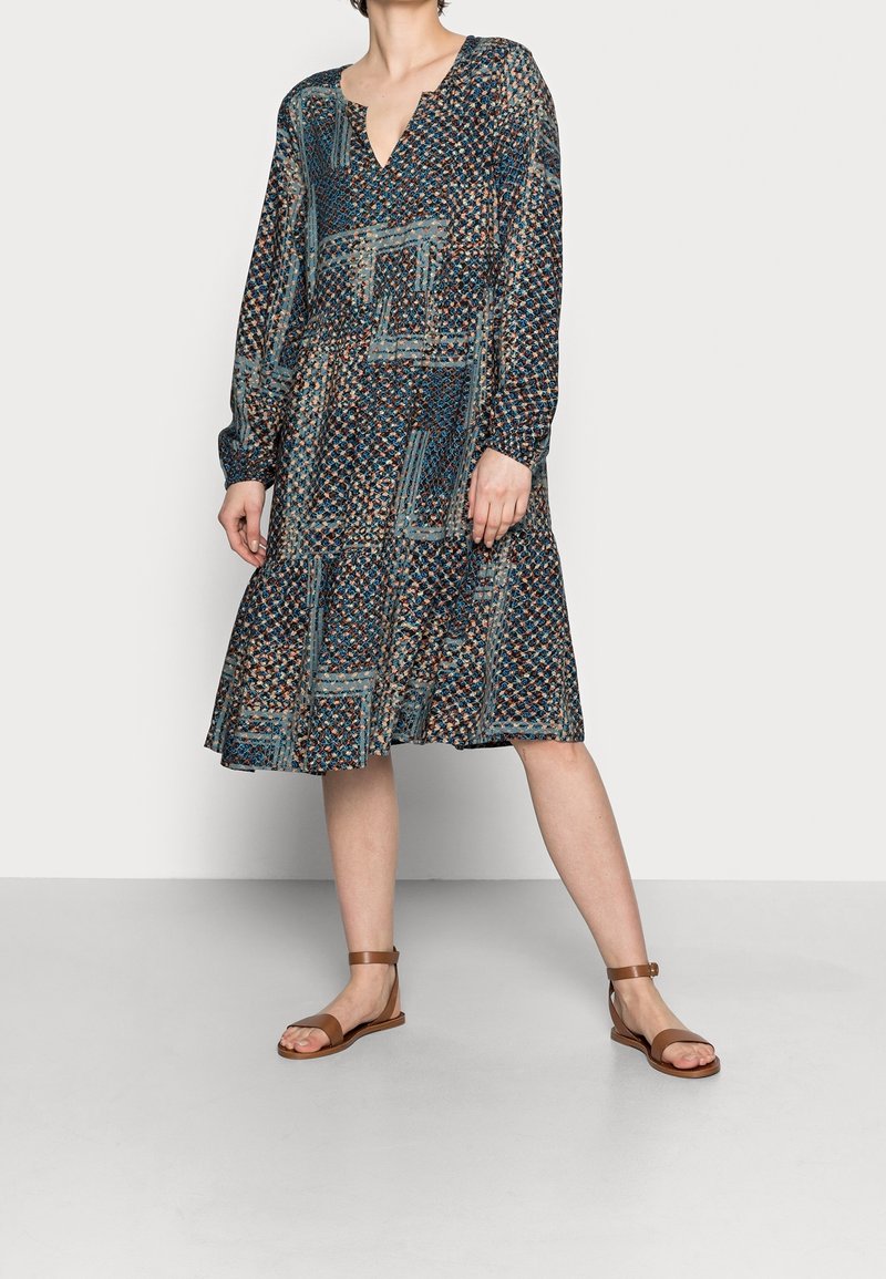 Femme portant une robe midi à manches longues avec un motif, un décolleté en V et des sandales plates marron, debout devant un fond uni.