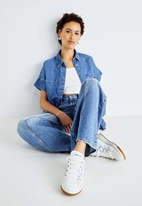 Zwei-teiliges Denim-Outfit bestehend aus einem hellblauen, verkürzten Hemd mit kurzen Ärmeln und hoch sitzenden Jeans. Kombiniert mit weißen Sneakers.