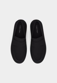 Mustade ümmarguste varvastega ja lamedate talladega Calvin Kleini libisevad mokassiinid, vaadatuna ülalt valgel taustal.