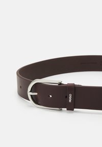 Cintura in pelle marrone con una fibbia in metallo argentato. Presenta una superficie liscia e include fori per la regolazione. Il logo del marchio è stampato in bianco.