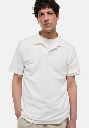 Homme aux cheveux bouclés et foncés portant un polo blanc à manches courtes par-dessus un sous-vêtement blanc, debout avec une main dans la poche d’un pantalon beige.