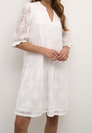 Femme portant une robe blanche en dentelle avec des motifs floraux, des manches trois-quarts bouffantes et un décolleté en V, debout devant un fond uni.