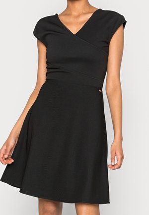 Woman wearing a black short-sleeve dress with a V-neck and fitted waist, standing against a plain light background.