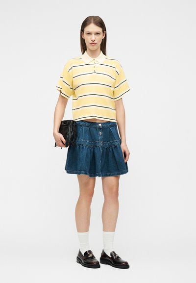 Γυναίκα που φοράει κίτρινη ριγέ πόλο μπλούζα, μπλε τζιν φούστα, λευκές κάλτσες, μαύρα μοκασίνια, κρατώντας μια μαύρη τσάντα φάκελο με κρόσσια, στέκεται μπροστά σε λευκό φόντο.