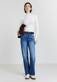 Eine Frau steht und hält eine braune Jacke, trägt ein weißes Langarmshirt, hochtaillierte blaue Jeans, einen braunen Gürtel und weiße Sneakers vor einem schlichten Hintergrund.