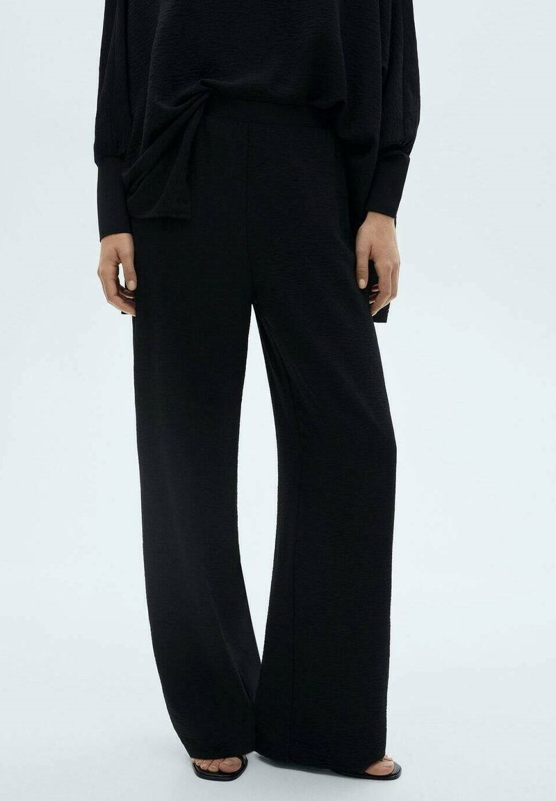 Black wide-leg pants made of textured fabric, featuring a high waist and minimalistic design. Accented with a side tie detail.