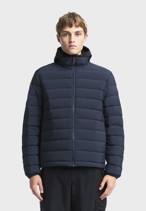 Jeune homme portant une doudoune matelassée bleu marine avec un col montant et une fermeture éclair frontale, debout devant un fond clair uni.