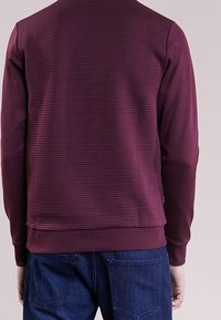 Sudadera de cuello redondo burdeos con textura acanalada, mangas largas y un diseño liso en la parte trasera, combinada con jeans de mezclilla azules.