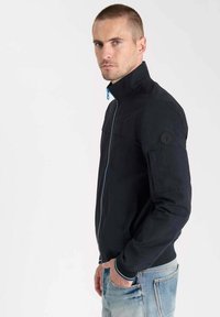 Veste noire avec un col haut, fermeture éclair bleue et un écusson sur la manche gauche ; associée à un jean bleu clair. Texture lisse, design moderne.