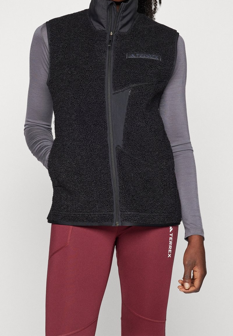 Black fleece vest with high collar, front zipper, and logo detail. Worn over a long-sleeve gray top and paired with maroon fitted pants.