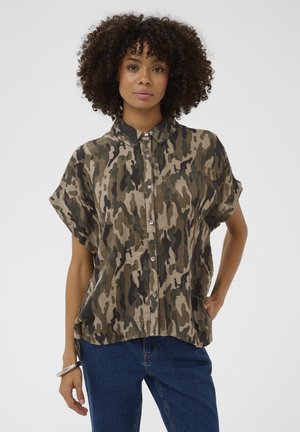 Žena s kučeravými vlasmi, oblečená v maskáčovej košeli s krátkym rukávom a modrých džínsoch, stojaca s jednou rukou v vrecku a neutrálna výrazom tváre.