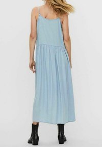 Vaaleansininen farkkumaximekko, jossa on säädettävät olkaimet, kerätty vyötärö ja virtaava siluetti; yhdistetty mustiin nilkkasaappaisiin.