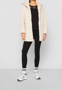 Manteau en fausse fourrure crème avec capuche, superposé à un haut noir à dentelle, associé à des leggings noirs et des chaussures de sport blanches avec des accents.