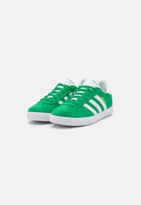 Green suede sneakers with white stripes, textured rubber sole, round toe, and white laces. Gold logo detail on the side.