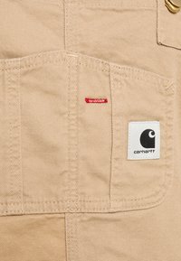 Béžová pracovní látka s kapsou a červeným prošíváním. Obsahuje černobílou nášivku s logem Carhartt na kapse.