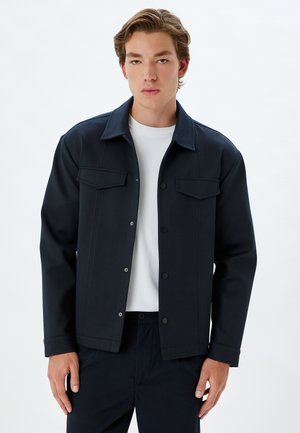 Chaqueta azul marino hecha de tela gruesa, con cuello, dos bolsillos en el pecho y cierre de botones. Lleva puesta una camisa blanca.