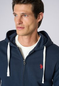 Felpa con zip di colore navy, con cordini bianchi, presenta un piccolo logo rosso sul lato sinistro del petto, realizzata in morbido cotone.