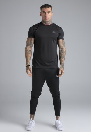 Tetoválásokkal rendelkező férfi formás fekete pólóban, fekete, logókkal díszített melegítőnadrágban és fehér sportcipőben szürke háttér előtt áll.