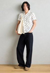 Short-sleeve, white button-up shirt with dark patterns, paired with loose-fitting dark pants and beige shoes. Holds a cream-colored tote bag.