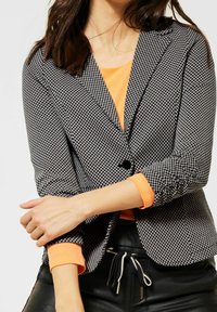 Sako s černobílým vzorem houndstooth s jednočtvrtečním zapínáním, s ohrnutými rukávy přes oranžový top a černé kalhoty.