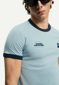Light blue short-sleeve t-shirt with navy blue collar and cuffs. Features printed text: "quality performer" and logo on the chest.