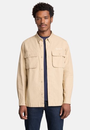 Junger Mann, der ein beiges Hemd über einem dunkelblauen T-Shirt und dunkelblauen Jeans trägt, vor einem schlichten hellen Hintergrund stehend.