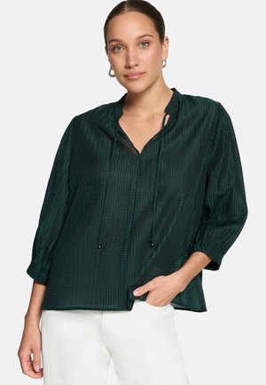 Femme en blouse texturée vert foncé à manches trois-quarts et pantalon blanc, posant avec une main dans la poche devant un fond uni.