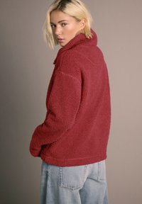 Femeie blondă purtând o jachetă roșie texturată din fleece și blugi bleu deschis, privind peste umăr pe un fundal bej.