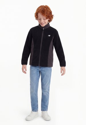 Jonge roodharige jongen staat, draagt een zwart jack met rits, lichtblauwe jeans en witte sneakers, glimlacht met ogen naar beneden gericht.